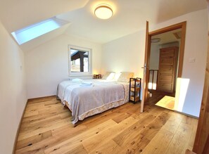 Un dormitorio con tragaluz, piso de madera, cama con ropa de cama, mesita de noche con lámpara y armario de madera.