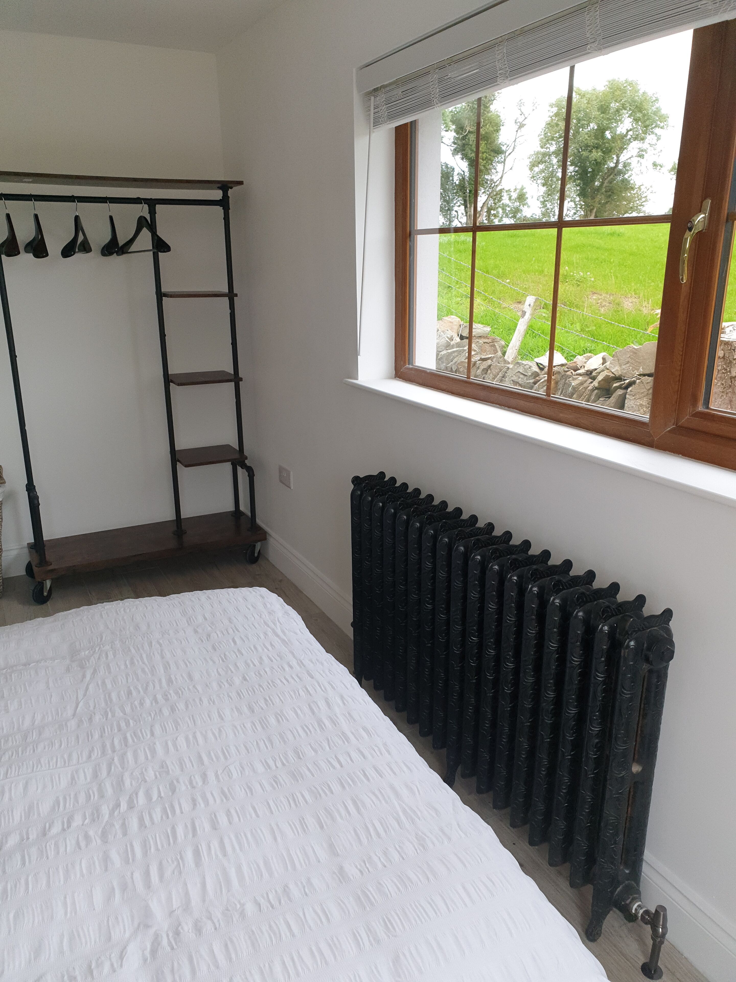 Un dormitorio con cama, radiador, ventana con vista a un campo verde y un perchero con ganchos.