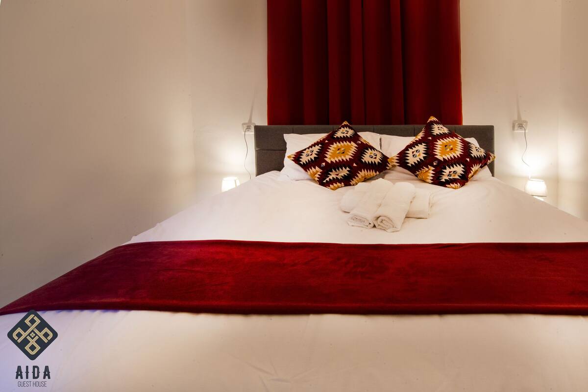 A neatly made bed with white linens and two patterned pillows, a red throw blanket, and a wall-mounted night light.