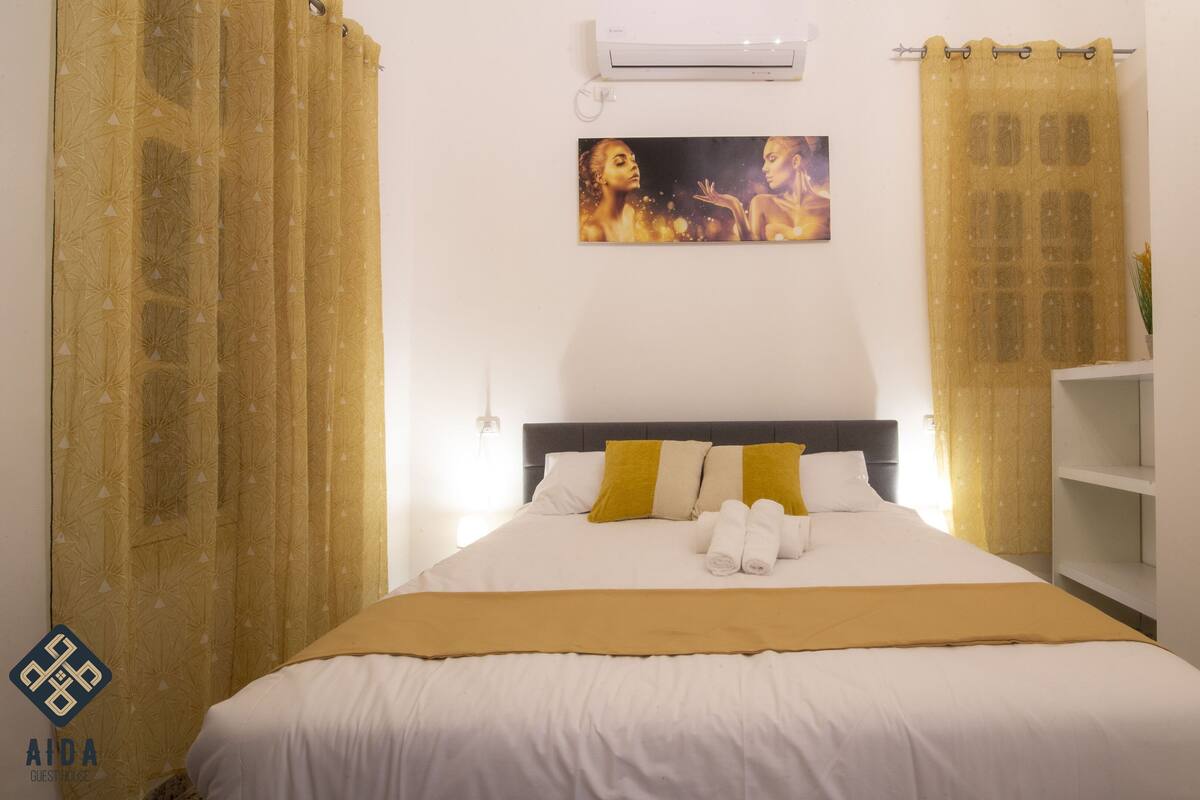 A hotel room with a bed, a painting above the bed, and a shelf to the right.