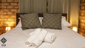 A neatly made bed with patterned pillows and rolled white towels, set against a brick wall with a curtain and a wall-mounted lamp. - Aida Guest House (Nazareth)