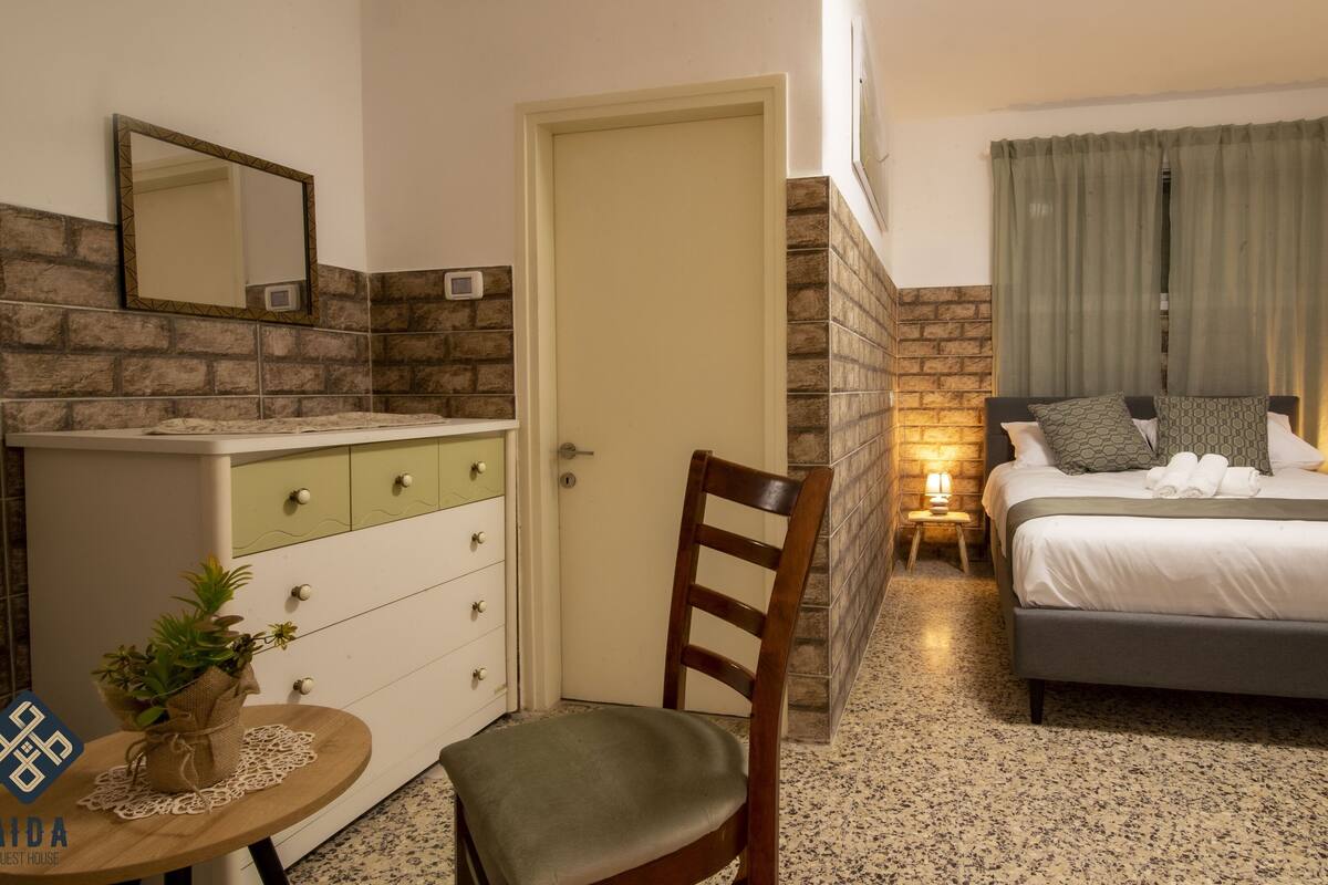 A hotel room with a bed, a chair, a dresser, a small table with a plant, and a mirror.