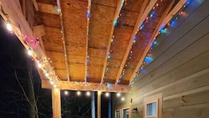 Una terraza de madera con barandilla, dos sillas, una mesa pequeña y luces colgantes.