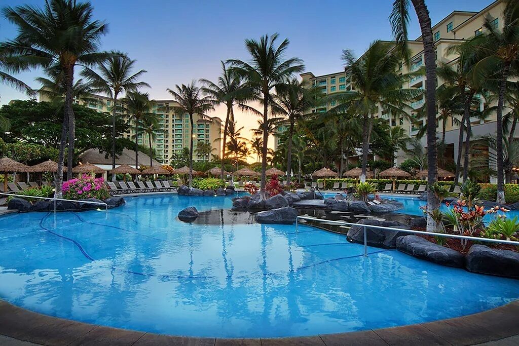 A resort pool with lounge chairs, palm trees, and a building in the background.