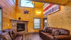 L’intérieur chaleureux d’un chalet avec une cheminée en pierre, des canapés en cuir, des murs en bois et une grande fenêtre.