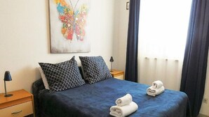 Ein ordentlich bezogenes Bett mit dunkelblauer Bettwäsche, zwei gemusterten Kissen und zwei aufgerollten Handtüchern.