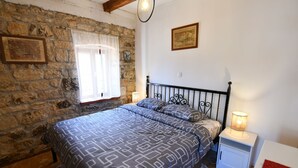 A bedroom with a stone wall, a bed with patterned bedding, a nightstand with a lamp, and a framed picture on the wall. - Apartment Rural Podastrana (Sinj)