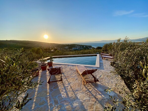 Zona junto a la piscina con sillones de descanso y plantas en macetas, con vistas a un paisaje pintoresco con colinas y una masa de agua.