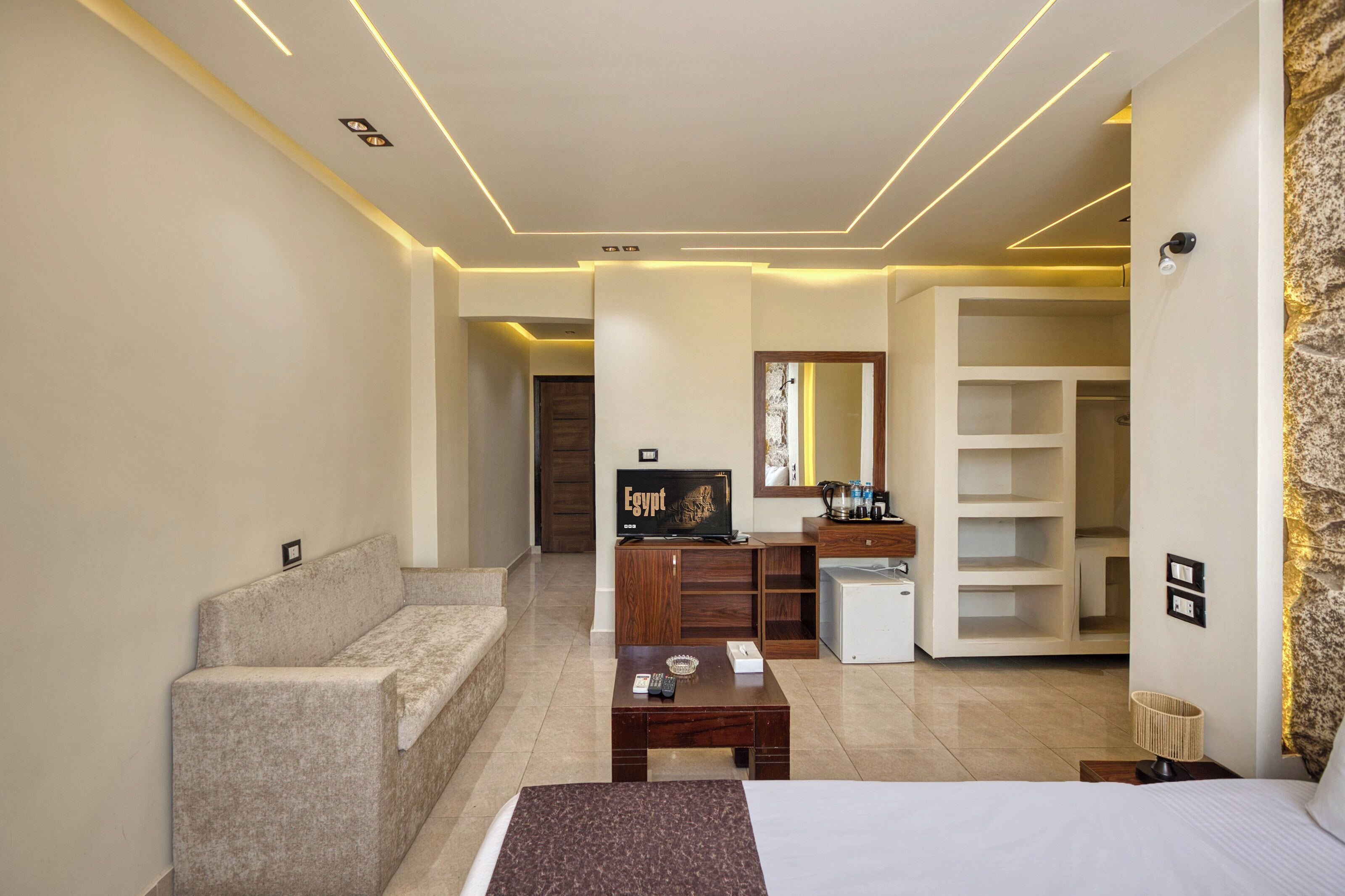 A modern hotel room with a bed, a sofa, a desk, a television, and a minibar.