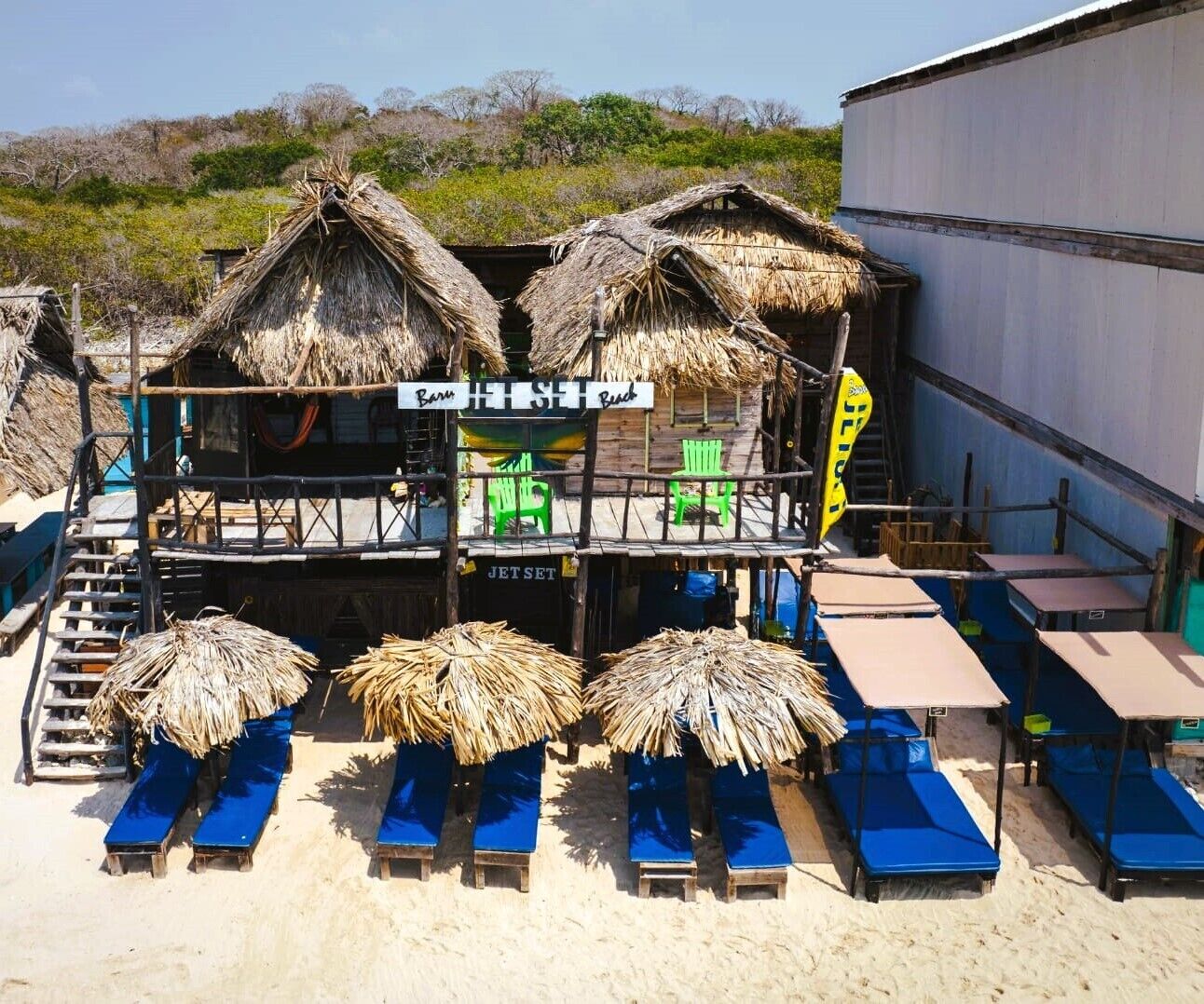 Ein Gebäude am Strand mit Reetdächern, einem Jetset Schild und blauen Liegestühlen.