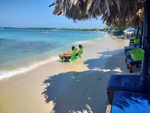 Une plage où deux personnes sont assises sur des chaises vertes, sous un toit de chaume, face à la mer.