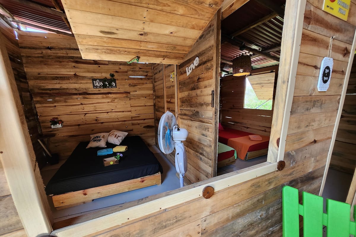 A wooden cabin with a bed, a fan, and a small table with items on it.