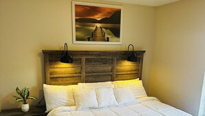 A bedroom with a wooden headboard, a large bed with white bedding, a nightstand with a plant, and a painting of a sunset over water on the wall. - The Great Escape, King suite, Kid friendly (Sevierville)