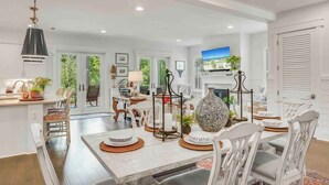 Un comedor amplio con una mesa blanca dispuesta para seis personas, rodeada de sillas blancas. Un jarrón decorativo y un candelabro están sobre la mesa.