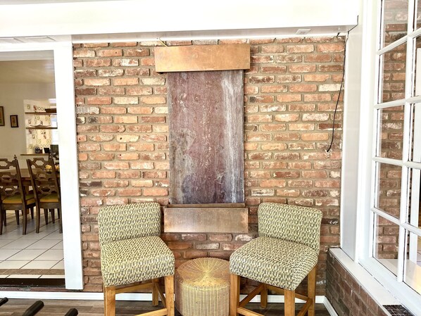 Een rustiek interieur met een bakstenen muur, twee gestreepte fauteuils, een geweven poef en een houten plank.
