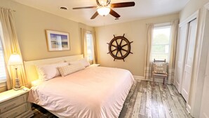 Ein Schlafzimmer mit einem Bett, einem Deckenventilator, einem Steuerrad, einem Sessel und einem Fenster mit Vorhängen.