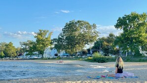 Ein Strand mit einer Person, die auf einer Decke sitzt, Bäumen und Häusern im Hintergrund.