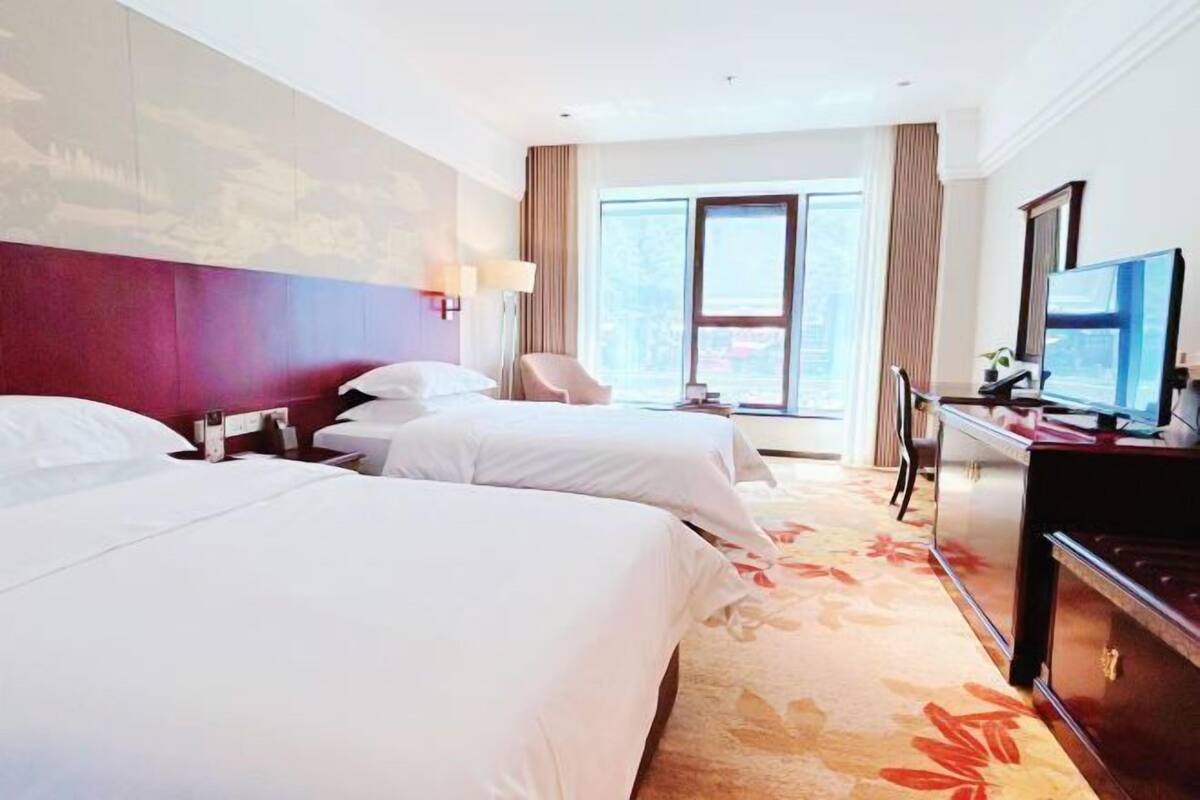 A hotel room with two beds, a desk, a television, and a large window.