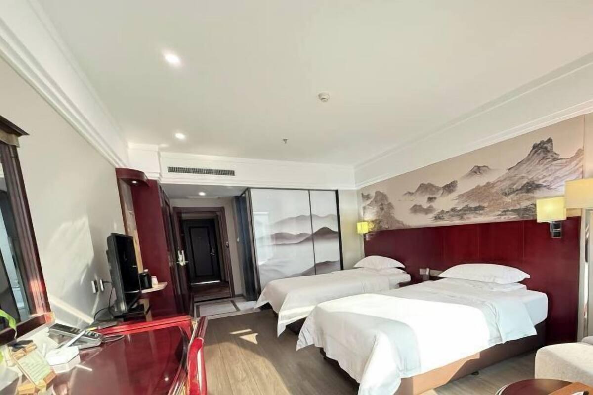 A hotel room with two beds, a desk, a TV, and a painting on the wall.