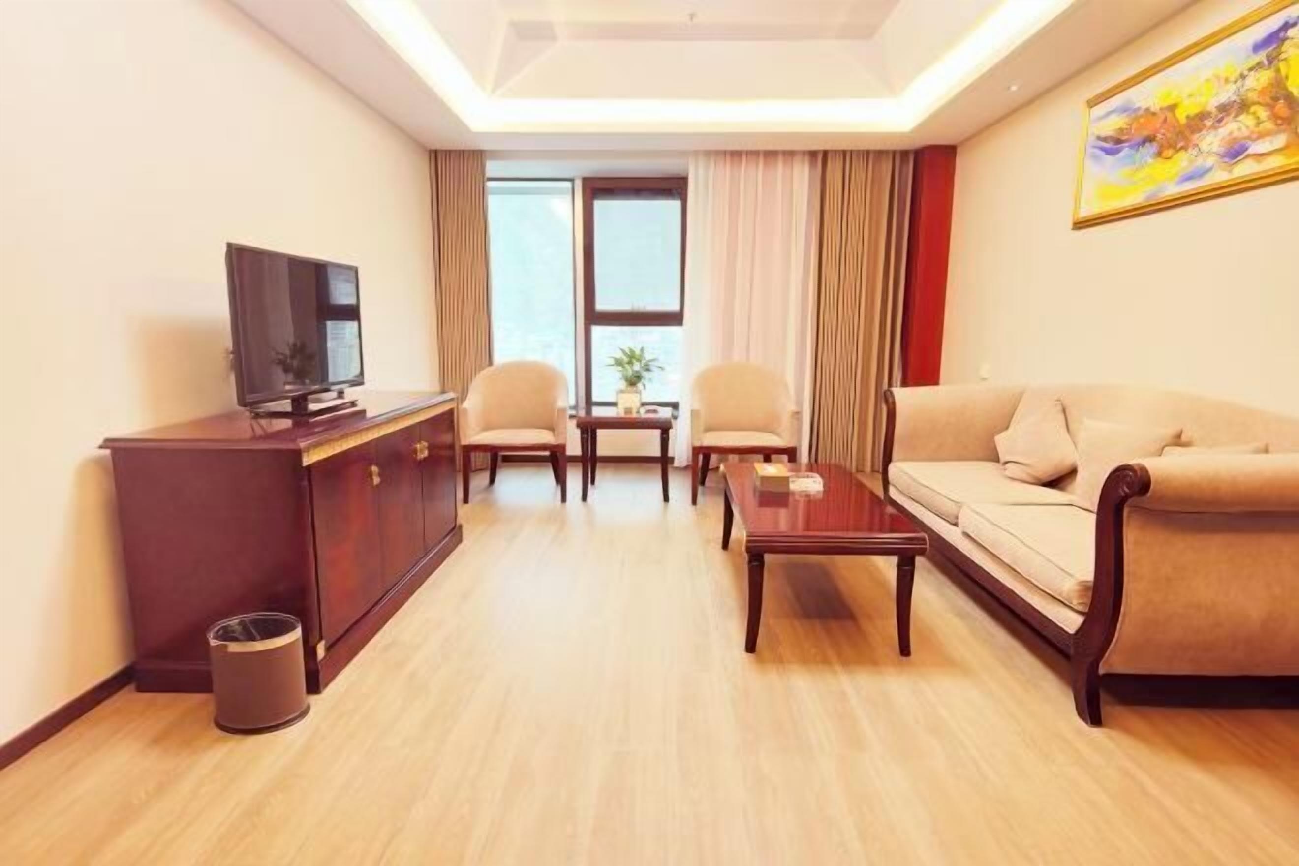 A hotel room with a sofa, a TV, a small table, two chairs, a cabinet, and a window with curtains.