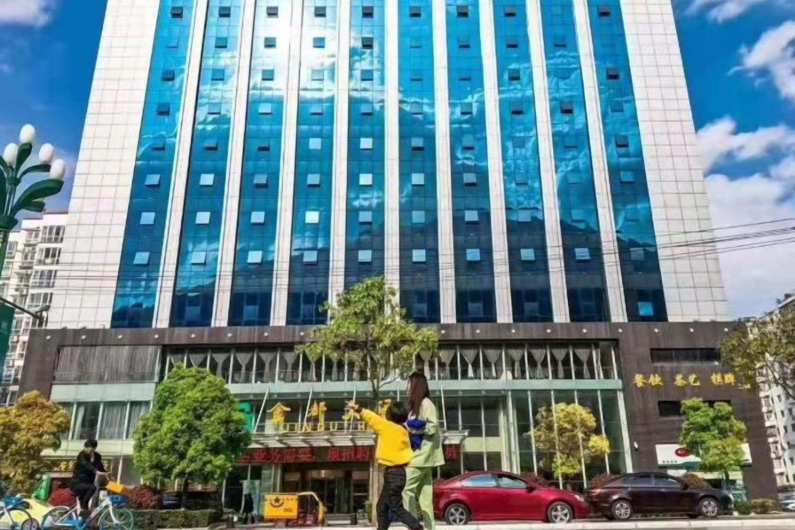 A tall building with a modern glass facade, a ground-level entrance with a sign, and a few cars parked in front.