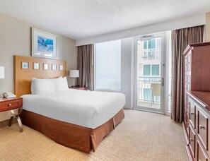 A hotel room with a large bed, a wooden headboard, a desk with a telephone, a nightstand with a lamp, a large window with curtains, and a balcony with a chair. - Hilton Ocean Beach Club Oceanfront Resort 1 bedroom with Oceanview balcony! (Virginia Beach)