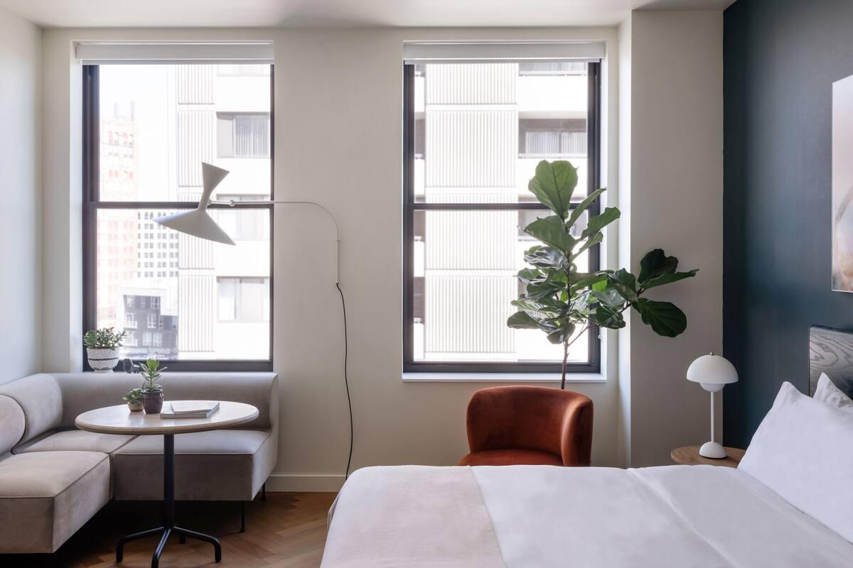A modern hotel room with a bed, a sofa, a small table, a potted plant, and large windows offering a view of the city.