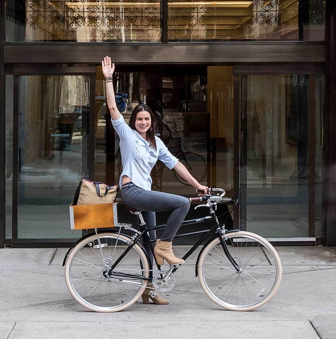 A person sitting on a bicycle with a bag attached to the back, in front of a building with large windows and a decorative metal railing.