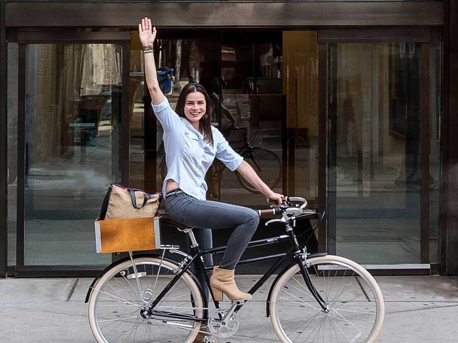 A person sitting on a bicycle with a bag attached to the back, in front of a building with large windows and a decorative metal railing.