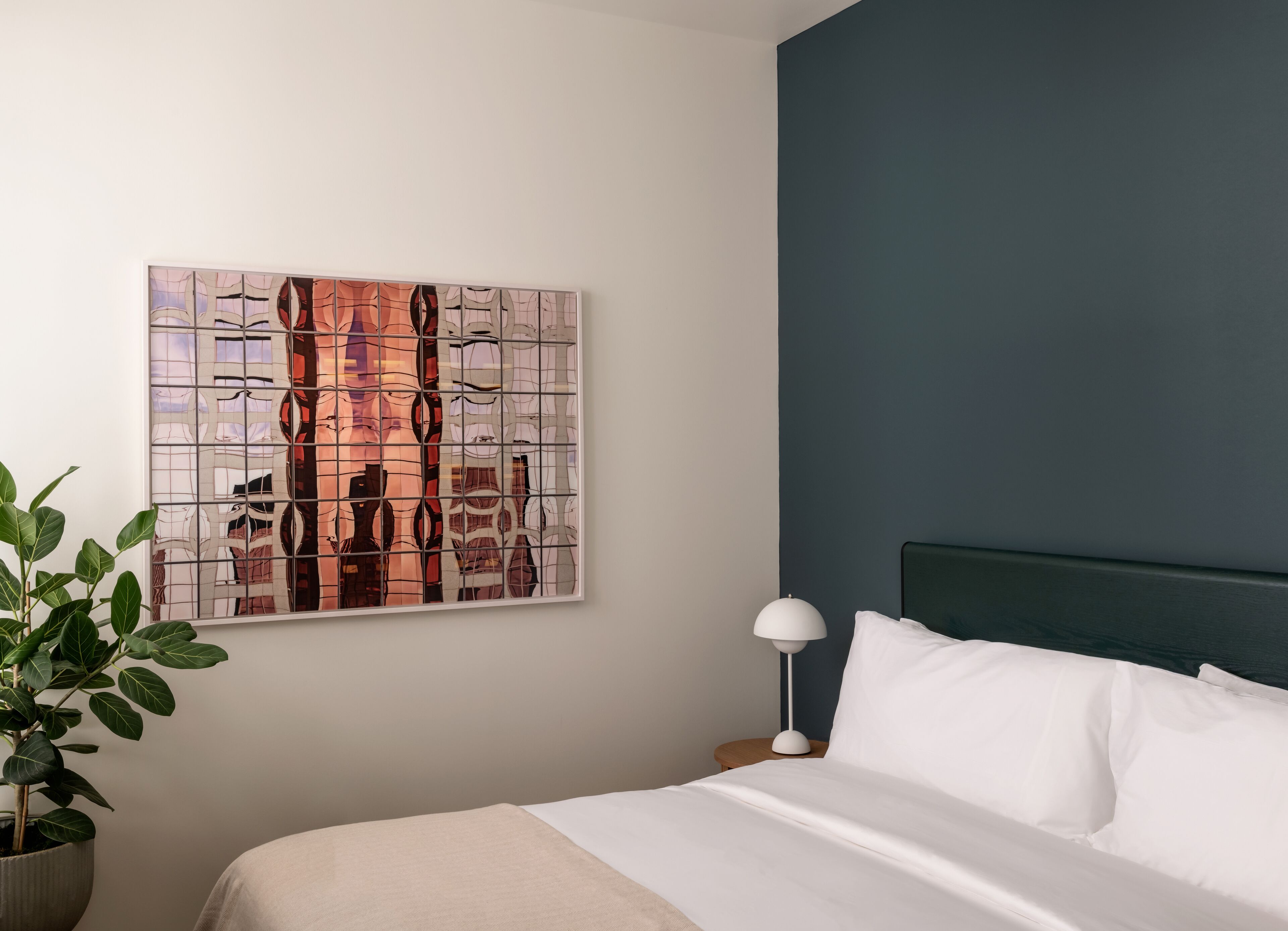 A hotel room with a bed, a nightstand, a lamp, a potted plant, and a wall art piece.