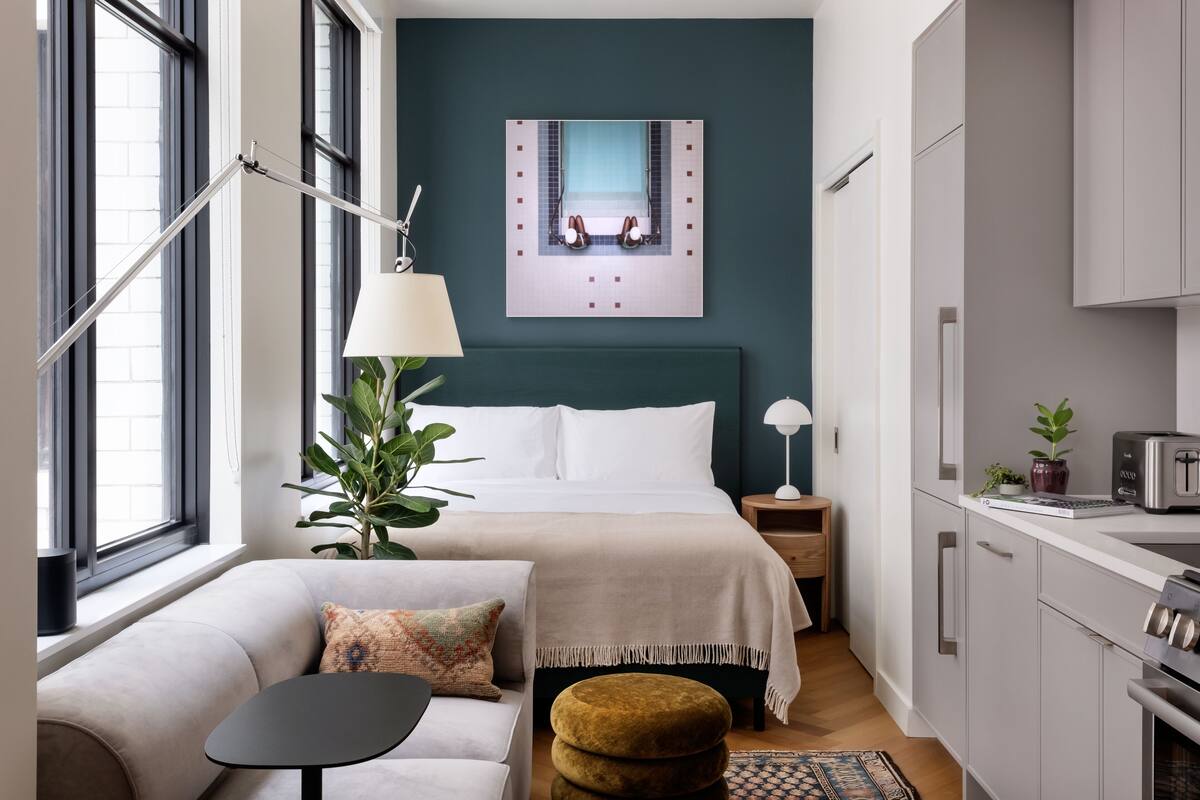 A compact hotel room with a bed, bedside tables, a small sofa, a dining area, and a kitchenette.