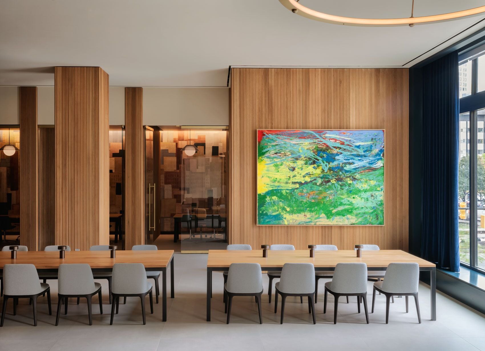 A modern conference room with a large wooden table, grey chairs, and a colorful abstract painting on the wall.