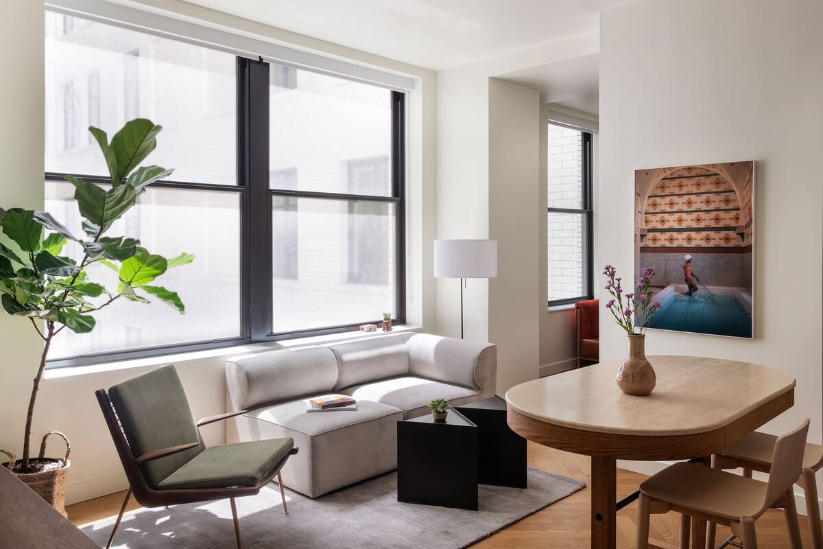 A modern living room with a sofa, a coffee table, a dining table with chairs, a lamp, and a large window with blinds.