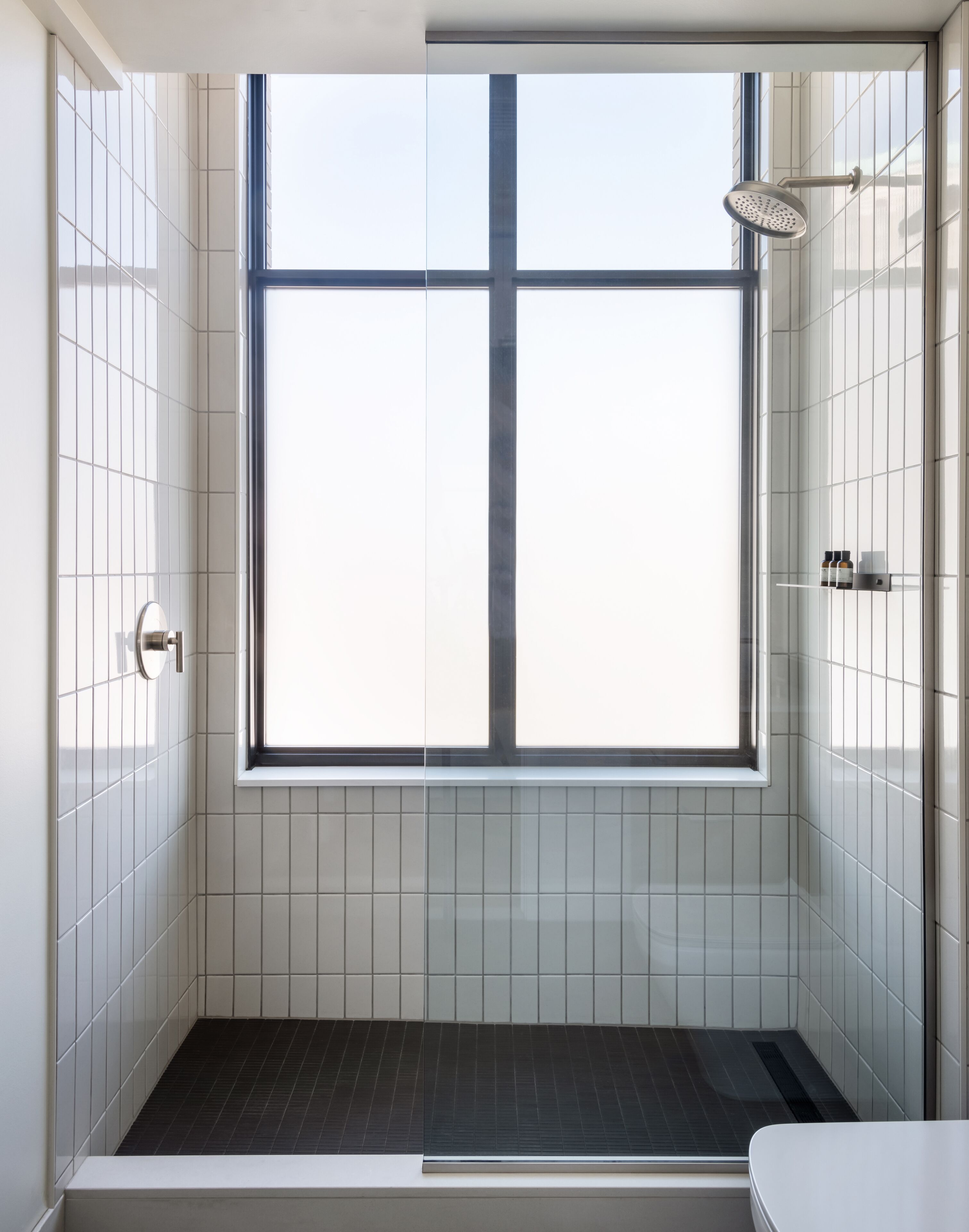 A modern shower with a large window, white tiled walls, and a dark floor.