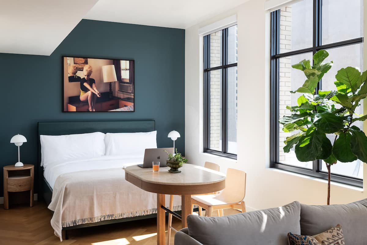 A modern hotel room with a large bed, a round dining table, a sofa, and a large window with a view of the city.