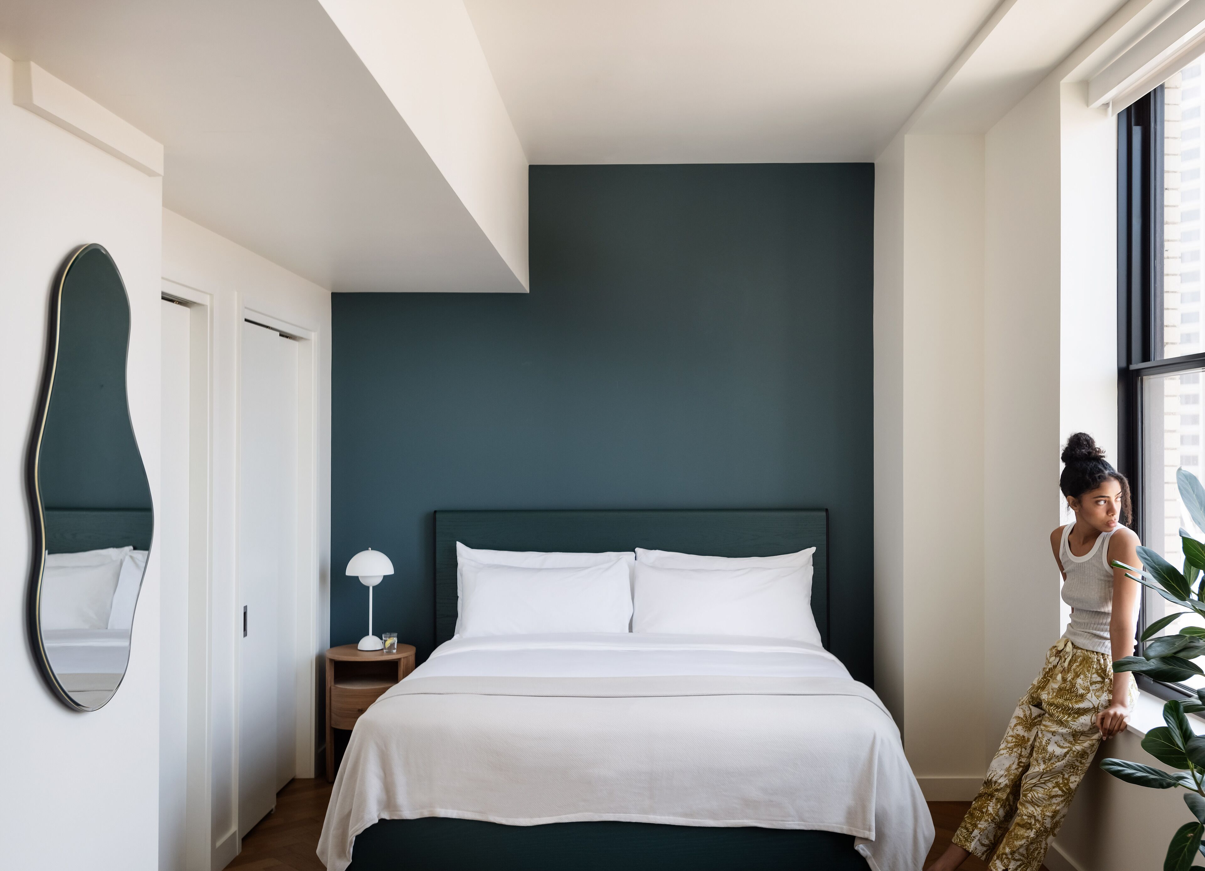 A modern bedroom with a large bed, a bedside lamp, a mirror, and a window with a view of buildings.