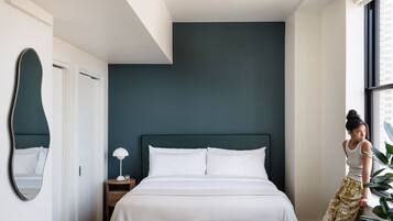 A modern bedroom with a large bed, a bedside lamp, a mirror, and a window with a view of buildings.