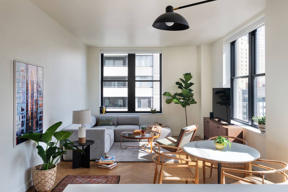 A modern living room with a sofa, coffee table, and dining area. There is a large window with blinds, a potted plant, and a painting on the wall.