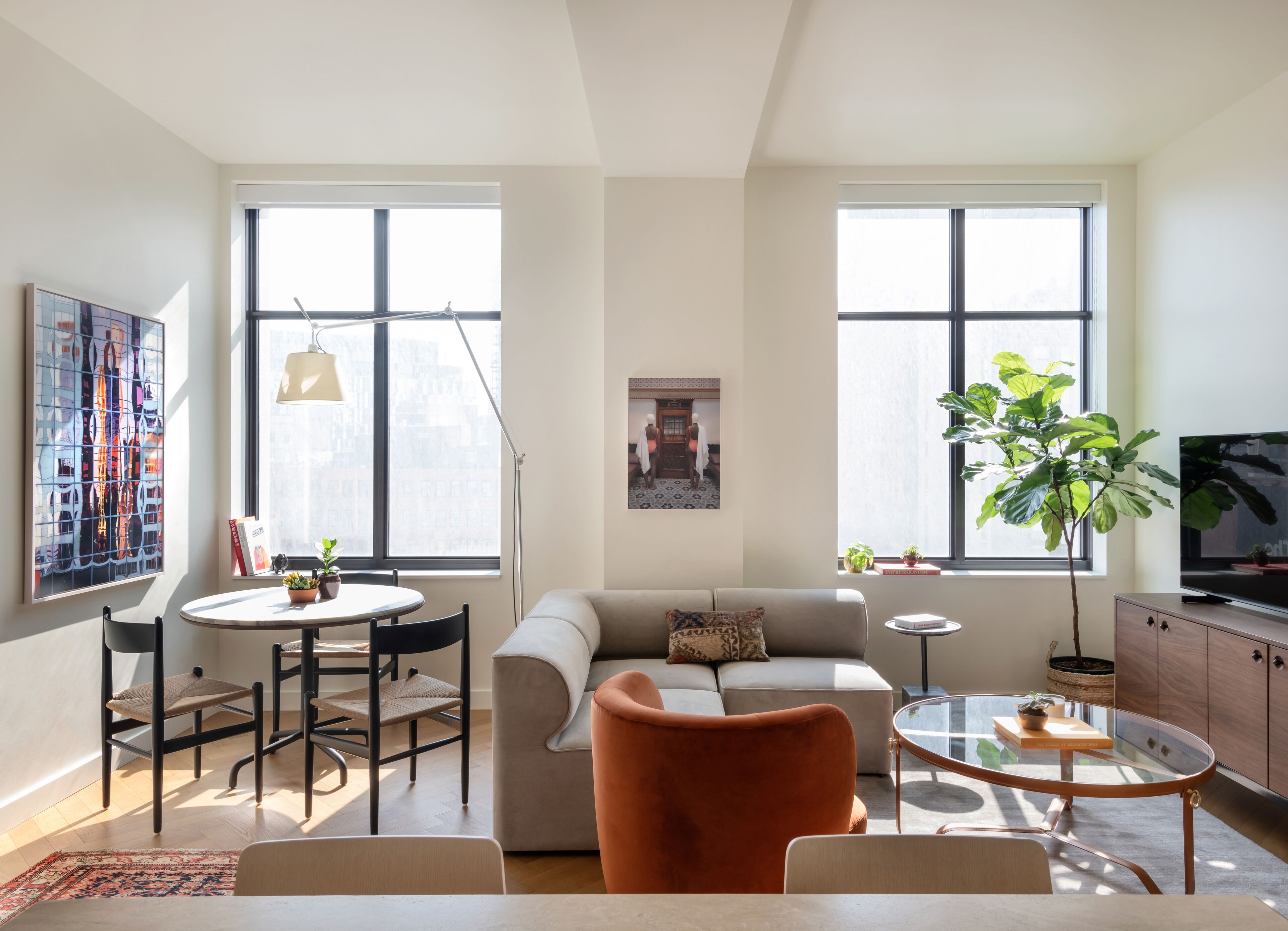 A modern living room with a sofa, coffee table, and a dining area with chairs and a small table. There are large windows providing natural light and a view of the city.