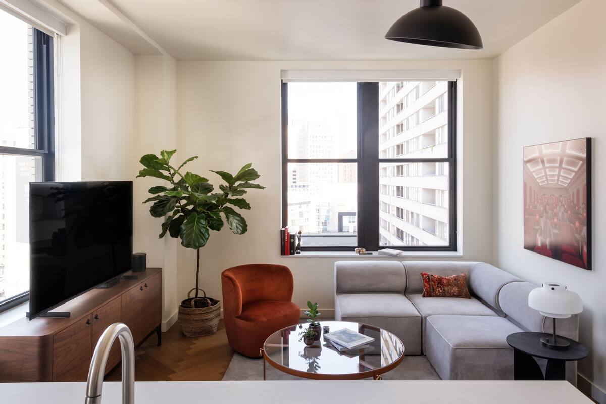 A modern living room with a flat-screen TV, a large potted plant, a red armchair, a grey sofa, and a round coffee table.