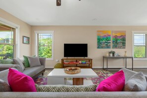 A living room with a sofa, coffee table, and TV. There are windows, a painting, and a small table with books and a plant.