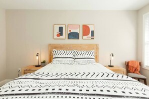 Quarto com cabeceira de madeira, colcha estampada, duas mesas de cabeceira com luminárias e arte abstrata na parede.