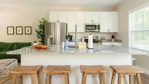 Eine moderne Küche mit weißen Schränken, Edelstahlgeräten und einer Arbeitsplatte aus Granit. Vier Holzbarhocker stehen an der Kücheninsel.