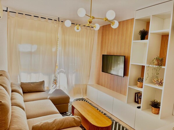 Un salon moderne avec un canapé beige, une table basse en bois, un téléviseur fixé sur un mur recouvert de panneaux en bois et une étagère blanche agrémentée de plantes.