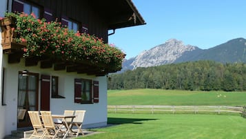 A chalet with a balcony adorned with red flowers, a dining area with wooden chairs, and a view of a mountain range.
