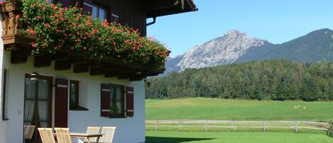 Ein Chalet mit einem Balkon, geschmückt mit roten Blumen, einem Essbereich mit Holzsesseln und Blick auf ein Gebirge.