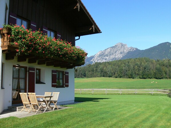A chalet with a balcony adorned with red flowers, a dining area with wooden chairs, and a view of a mountain range.
