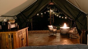 A tent with a wooden cabinet, two wicker chairs, a round table with a fire, and string lights.