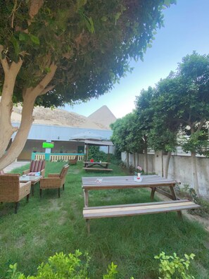 A grassy outdoor area with picnic tables and chairs, a wooden bench, and a large tree. - Villa Khufu Pyramids Hotel (Giza)