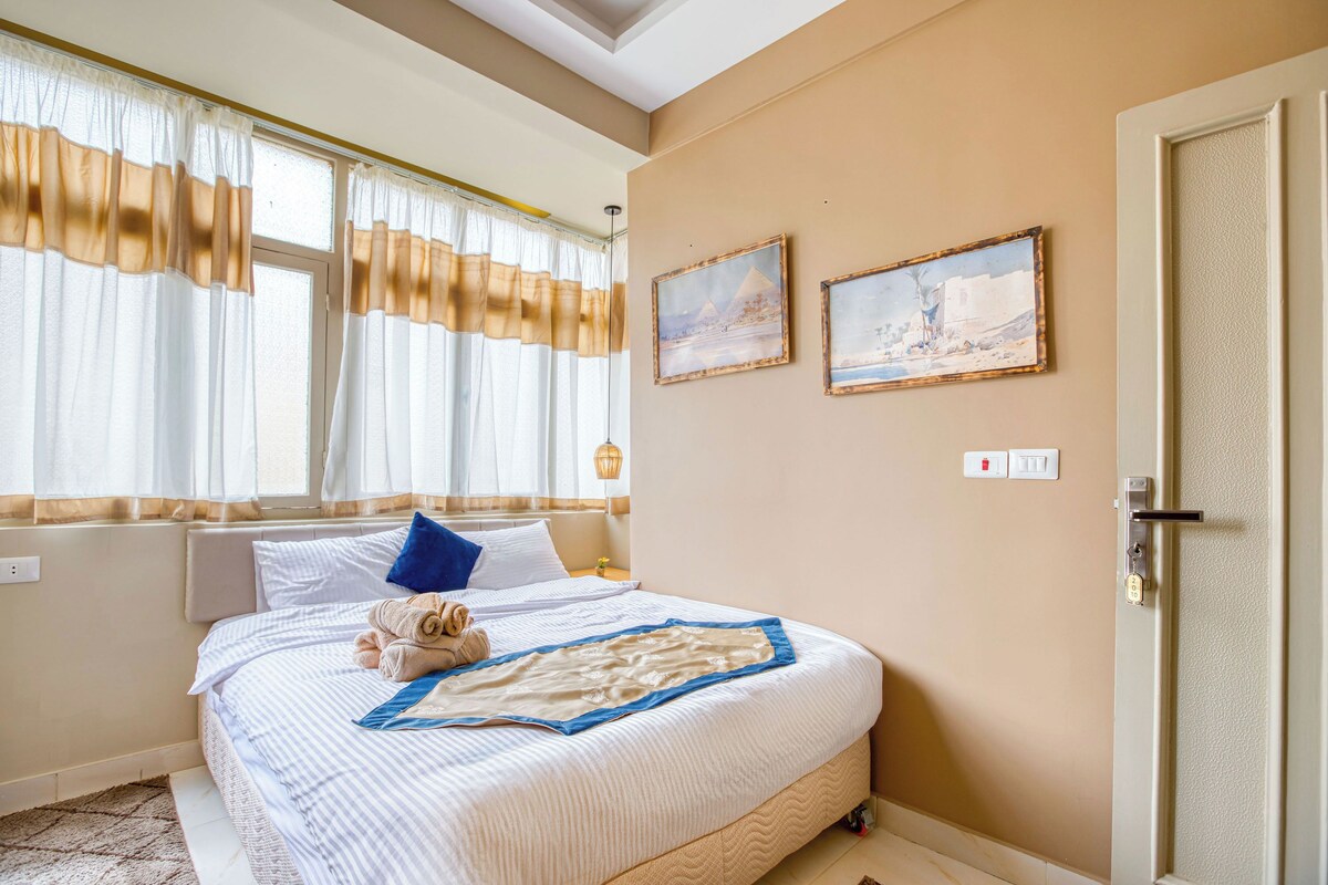 Una habitació d'hotel amb un llit, una finestra amb cortines, dos quadres al mur i una porta.